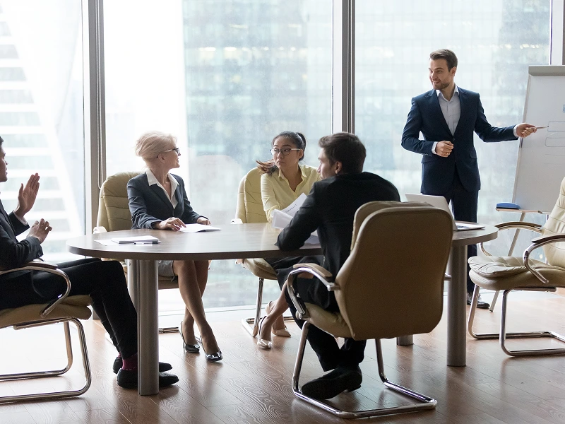 A man presents at a conference table with five colleagues seated, some taking notes. Large windows show a cityscape outside.