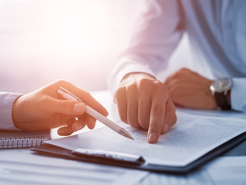 Two people sitting at a table reviewing a document. One hand holds a pen while the other points at the text. A notebook is on the side.